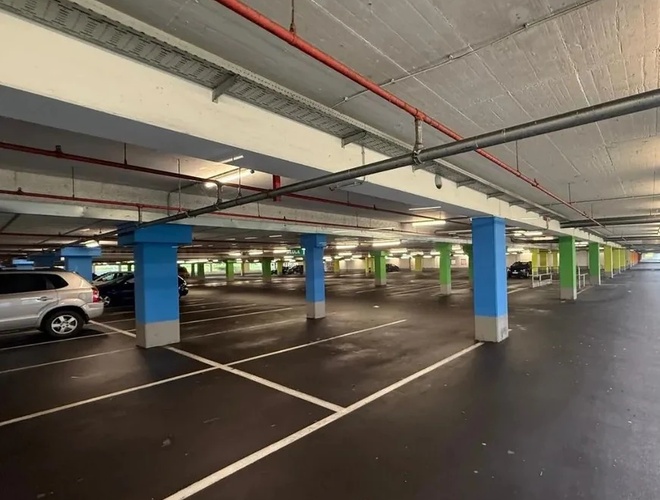 Podziemny parking z niebieskimi i zielonymi filarami, pustymi miejscami parkingowymi i kilkoma zaparkowanymi samochodami w tle.