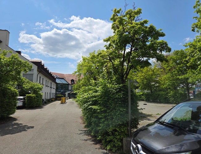 Atomis Hotel Munich Airport Parking – aparcamiento con árboles y un coche. Al lado hay un edificio verde. El área está bien cuidada y es espaciosa.