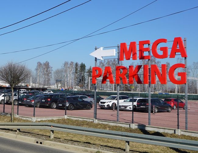 MEGA Parking - Terreno cercado, hay muchos coches. Gran cartel visible sobre el parking. Cielo despejado de fondo.