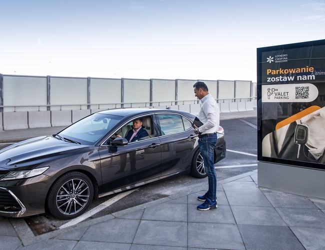Parkovanie pre osobné vozidlá – Letisko Chopina. Šofér v tmavom aute sa rozpráva s mužom. Vedľa je veľký informačný znak.