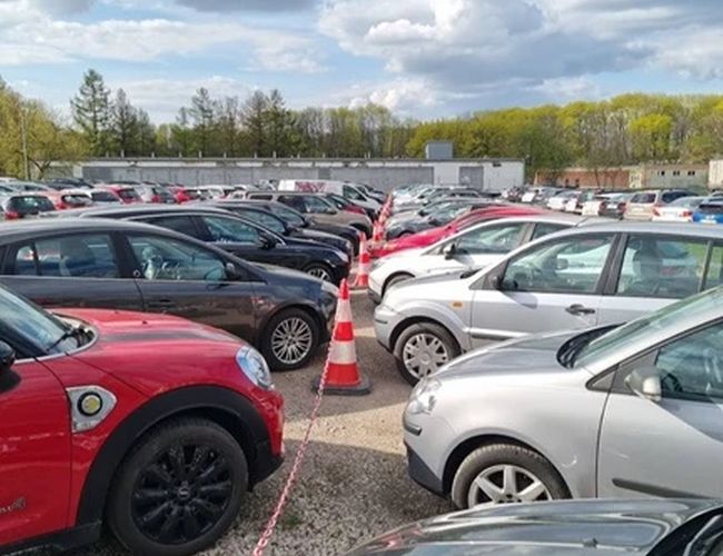 Parking Avanti - En la plaza de grava hay filas de coches. Conos marcan los caminos. Cielo parcialmente nublado de fondo.