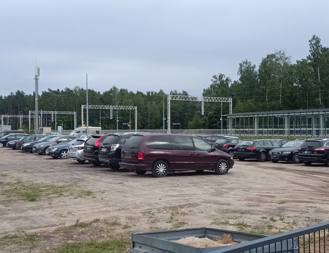 Smart parking - Parking jest nieutwardzony, otwarty i przestronny. Widać zaparkowane samochody osobowe. W tle znajdują się drzewa i budynek.