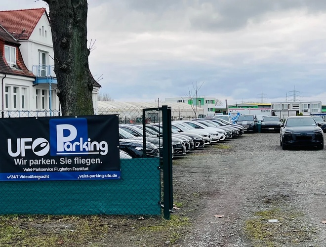UFO Valet Parking - Na pierwszym planie brama wjazdowa do parkingu. Widac zaparkowane prostopadle samochody. W tle widać drzewo oraz biały budynek.