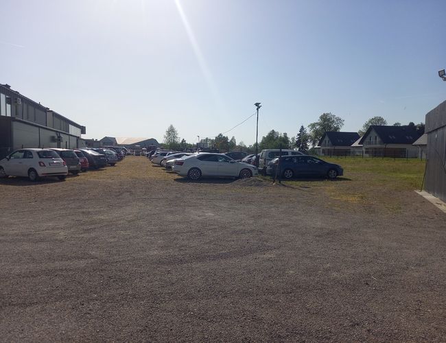 AutoParking24h - Parking zapełniony samochodami. Teren żwirowany, ogrodzony siatką. Po bokach stoją budynki mieszkalne.