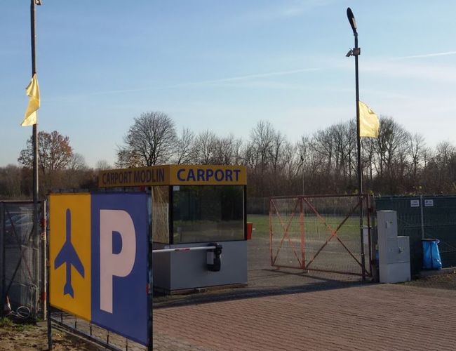 Parking Carport – Na zdjęciu jest ogrodzony obszar z bramą. Znajduje się na nim budka z napisem Carport Modlin. Na pierwszym planie jest znak parkingowy.