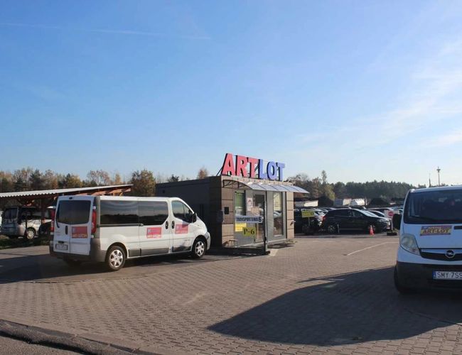 ArtLot 2 – En el aparcamiento se ven dos minibuses blancos. Al fondo están aparcados coches. El entorno es acristalado y se puede ver un pequeño edificio.