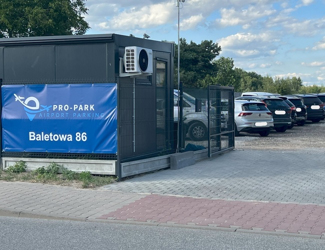 PRO-PARK – Parking z ogrodzeniem i tablicą informacyjną. Widoczne stojące samochody i mały budynek. Teren częściowo wyłożony kostką.