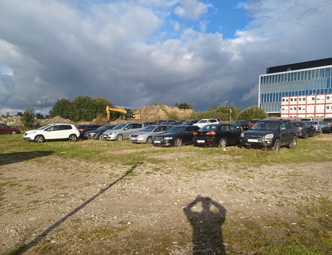 Maxi Parking – Estacionamiento en grava frente al edificio. Lleno de coches de varias marcas. Al fondo, una excavadora y árboles.