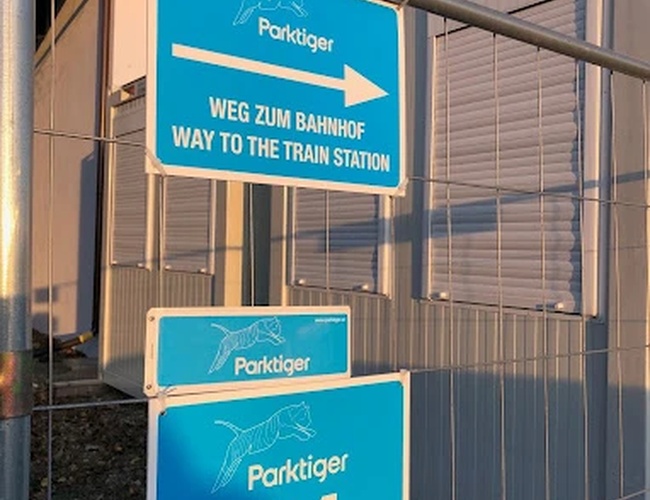 Parktiger Aeropuerto Estacionamiento 1 - Cercado con letreros azules informativos. Los letreros indican la dirección hacia la estación de tren. El contenedor está al lado.