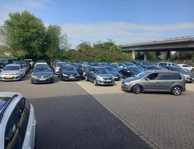 Parkovisko Elite Parken Köln - Parkovisko plné zaparkovaných áut. Doska pokrytá dlažbou, v pozadí most a zeleň.
