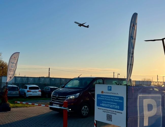 Parking Blisko Lotniska - Samochody stoją na ogrodzonym placu. Nad parkingiem leci samolot. Na płocie są rozwieszone flagi.