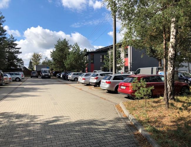 Parking Port - Na placu stoją zaparkowane samochody osobowe. Teren jest wyłożony kostką brukową i otoczony drzewami. Widać budynki w tle.