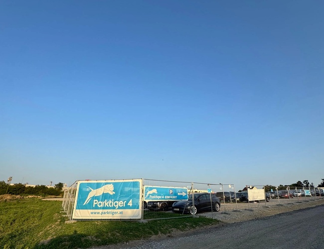 Amplio estacionamiento con un visible letrero azul con el nombre del estacionamiento Parktiger. Área cercada.