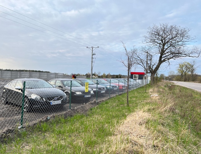 TRIP Parking - Parkovisko s oplotením z pletiva a autami. Územie susedí s asfaltovou cestou. Vedľa sa nachádzajú stromy.