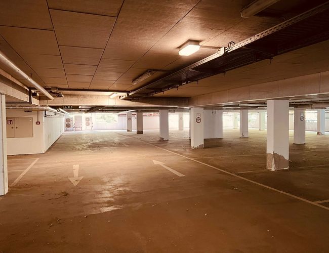 Sun Park Aeropuerto Garaje de Estacionamiento – Estacionamiento subterráneo con espacios vacíos. Interior iluminado y señalización en el suelo. Columnas blancas sostienen el techo.