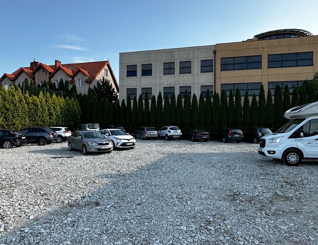 Parksist - Kamienné námestie s zaparkovanými autami. V pozadí domy a kancelárska budova. Nachádza sa tu aj rad vysokých ihličnanov.