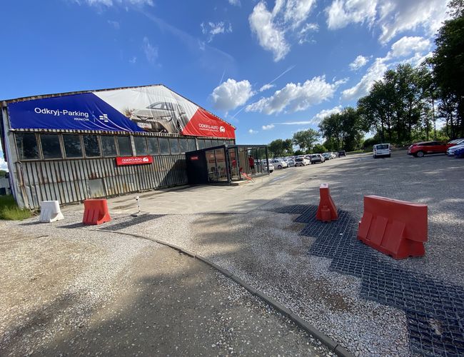 Odkryj-Parking - Budynek z dużą reklamą na ścianie. Obok plac z samochodami i barierkami. Na horyzoncie drzewa i niebo z chmurami.