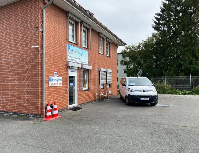Parking Parkservice Bremen Valet. Budynek administracyjny z czerwonej cegły z tabliczką informacyjną oraz busem transferowym zaparkowanym obok wejścia.