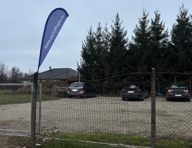 Wjazd na parking Park4flight Maramureș z otwartą metalową bramą, flagą informacyjną przy ogrodzeniu oraz kilkoma samochodami zaparkowanymi na przestronnym, utwardzonym placu w pobliżu lotniska.