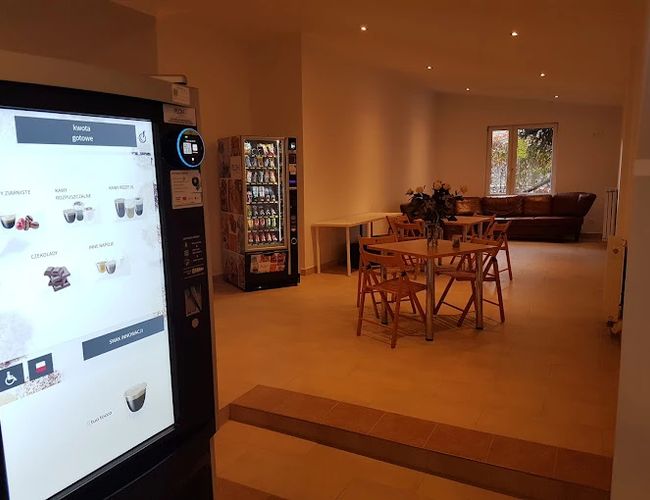 Parking Szyszkowa 39 – wewnętrzna poczekalnia z automatem do kawy. Stół z krzesłami oraz automat z przekąskami. Sofa przy oknie.