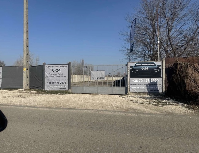 Puerta de entrada visible al aparcamiento con pancartas publicitarias. Área cercada, rodeada de árboles.