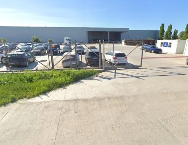 Szeroki wjazd na ogrodzony parking z otwartą bramą, na terenie widoczne liczne zaparkowane samochody, a w tle rozciąga się widok na morze oraz niską zabudowę przemysłową