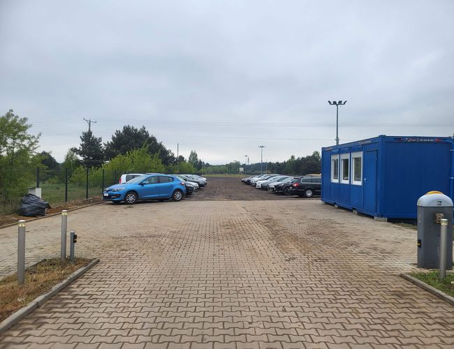 Primer Estacionamiento - En la plaza hay coches aparcados. Al lado hay un contenedor azul. El terreno está pavimentado con adoquines.
