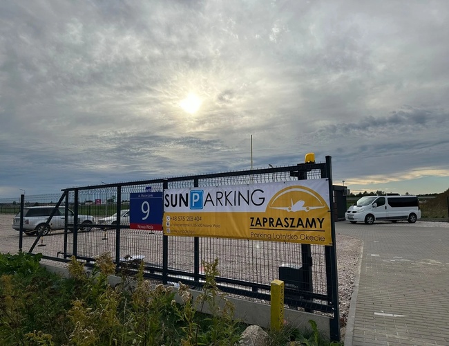 Parkovisko Sun - Na obrázku je viditeľné ohradené parkovisko. Na parkovisku je niekoľko áut. Vstup je ozdobený reklamným banerom.