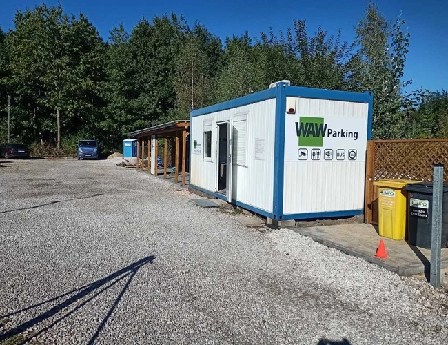 WAW Parkovanie - Štrkový pozemok s kancelárskym kontajnerom. Vedľa sú odpadkové koše. V pozadí sú vidieť autá a stromy.