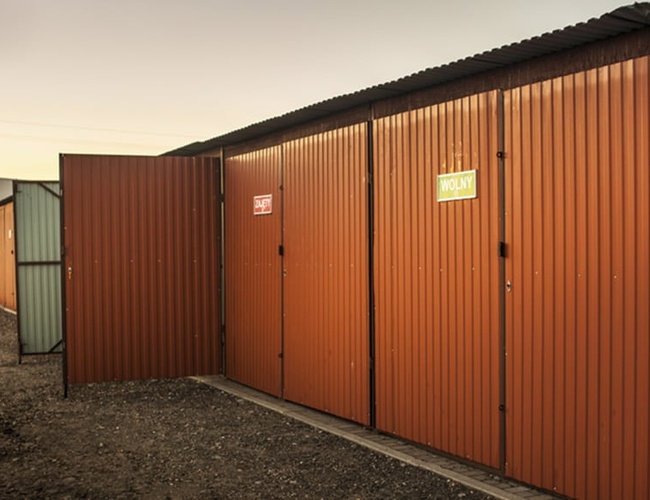 PERI Parking Garaże Premium – Kilka garaży z otwartymi drzwiami. Brązowe ściany, z napisami. Teren jest żwirowy i otwarty.