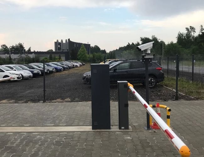 Parking LOT 24 - parking jest ogrodzony i monitorowany. W tle stoi szereg zaparkowanych samochodów. Brama wjazdowa jest opuszczona.