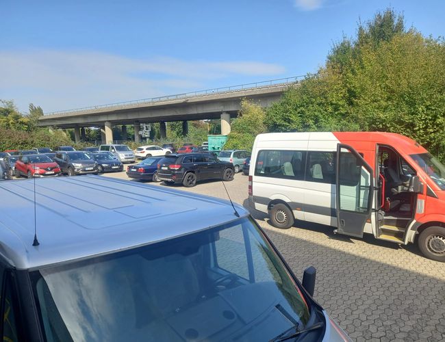 Parking Elite Parken Köln - Kilkanaście aut stoi na placu. Bus z otwartymi drzwiami widoczny jest z przodu. W tle widać wiadukt nad parkingiem.