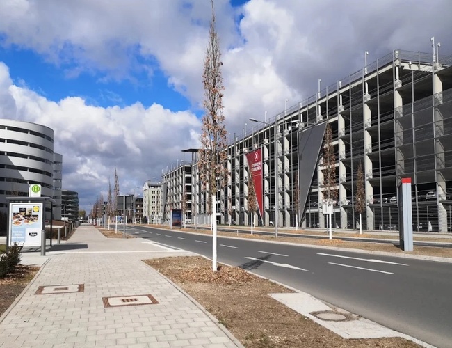 Wielopoziomowy parking P2 zlokalizowany przy terminalu lotniska, widok od strony ulicy z szerokim chodnikiem i drogą dojazdową.