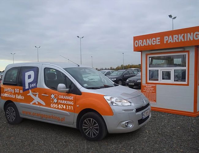 Orange Parking P3 - Samochód z oznaczeniem parkingu stoi obok budki. Teren jest żwirowy, w tle widoczne są latarnie i inne auta.