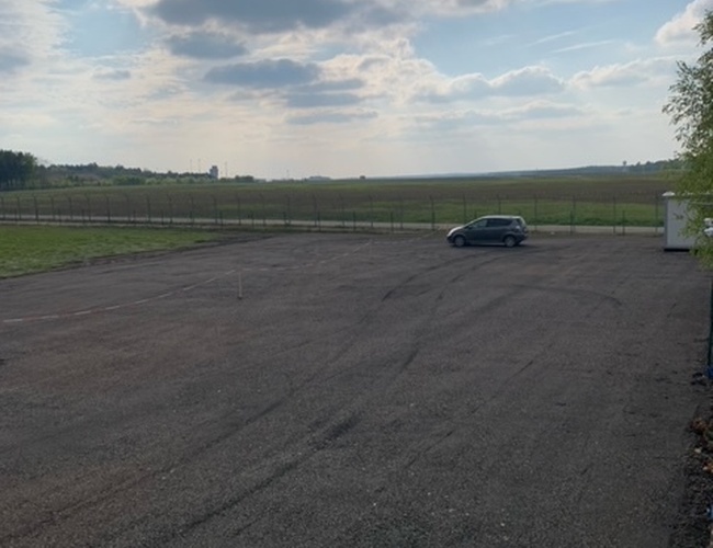 Estacionamiento con vista – un terreno vacío y de grava con un coche. En el fondo se ven campos y un cielo nublado. El área es abierta y espaciosa.