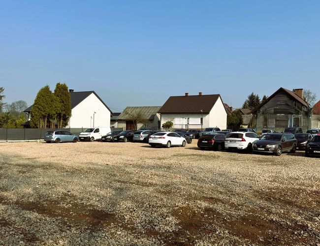 Parking Amber – Na żwirowym placu stoi wiele aut. W tle widać domy i drzewa.