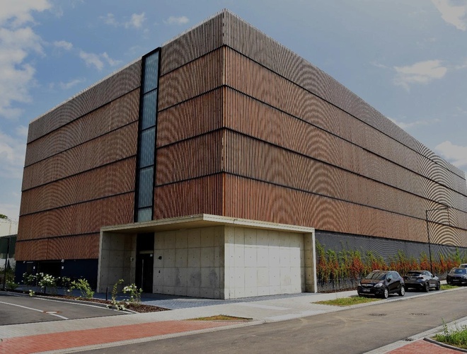 Parkhaus Flughafenparker – modernes Gebäude aus Holz und Beton. Davor stehen geparkte Autos. Nebenan wachsen grüne Sträucher.