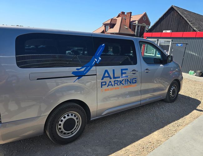 AleParking zadaszony - Srebrny bus z logiem parkingu stoi na terenie żwirowym. W tle są budynki i ogrodzenie.