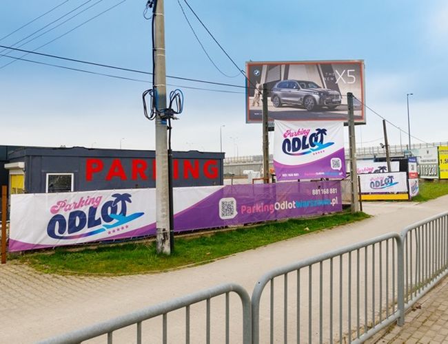 Parking Odlot – przestrzeń ogrodzona z reklamami na płotach. W tle znajduje się bilbord z samochodem. Obok widoczny jest chodnik.