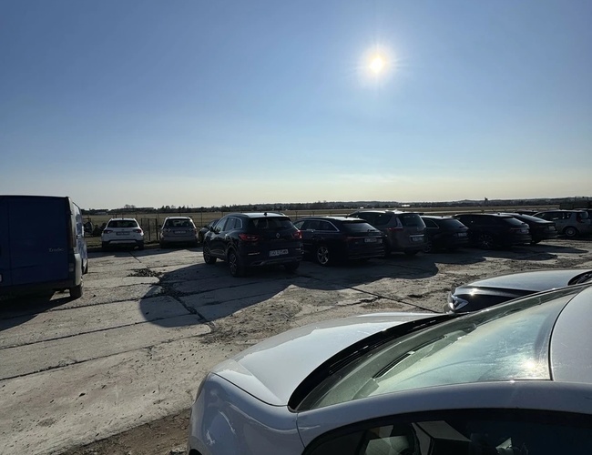 Parking MB Park – Na parkingu stoją samochody na żwirowej nawierzchni. Jest słoneczny dzień bez chmur.