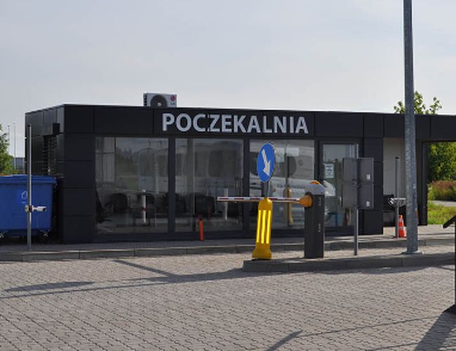 Parking Lotnisko – przed budynkiem poczekalni szlaban oraz znak drogowy. Budynek jest przeszklony, a w tle są drzewa.