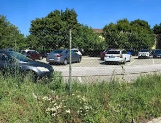 Ogrodzony parking z zaparkowanymi samochodami i budynkiem w tle, otoczony zielenią.