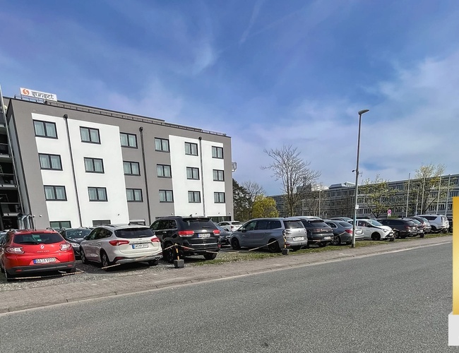 Parking Mein-Flughafenparkplatz-Frankfurt P3. Zaparkowane samochody wzdłuż ulicy obok bloku mieszkalnego, w tle jasne niebo.