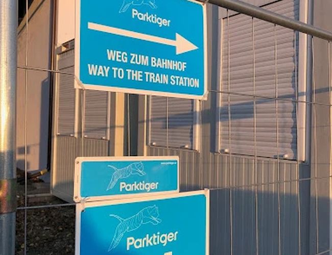 Parktiger Airport Parking 1 - Ogrodzenie z niebieskimi tablicami informacyjnymi. Tablice wskazują kierunek do stacji kolejowej. Kontener stoi obok.