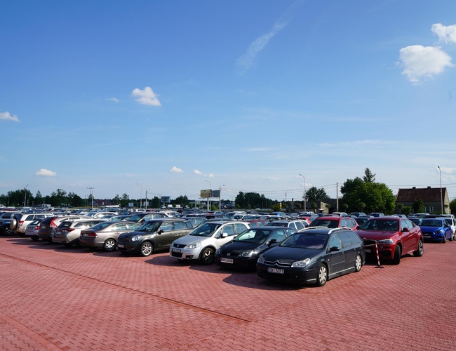 MEGA Parking - un gran espacio abierto lleno de coches. Los adoquines rojos cubren la superficie. A lo lejos se pueden ver árboles y edificios.