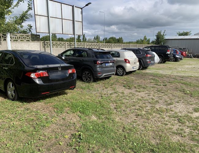 A1 Parking Max - Kilka samochodów stoi na placu. Teren jest ogrodzony betonowym płotem. Obok widać latarnię i drzewa.