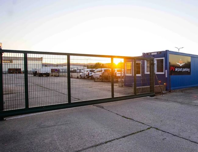 Wjazd na teren PV Airport Parking z oznaczeniem parkingu i budynkiem obsługi widocznym w tle, oświetlony światłem zachodzącego słońca.