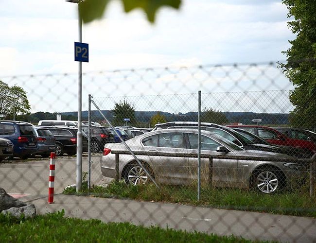 Parking z samochodami widziany przez ogrodzenie z siatki. W tle widoczne są kolejne miejsca postojowe oraz zieleń.