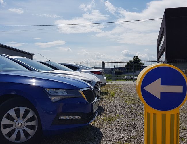 GOOD Parking - Na zdjęciu widoczne są zaparkowane samochody. Teren jest otwarty i nieogrodzony. Znak wskazuje kierunek jazdy.