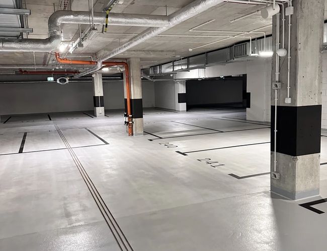 Supreme Parking Garage – Wnętrze pustego, podziemnego parkingu. Jasne oświetlenie i oznaczone miejsca postojowe. Betonowa konstrukcja z widocznymi rurami.